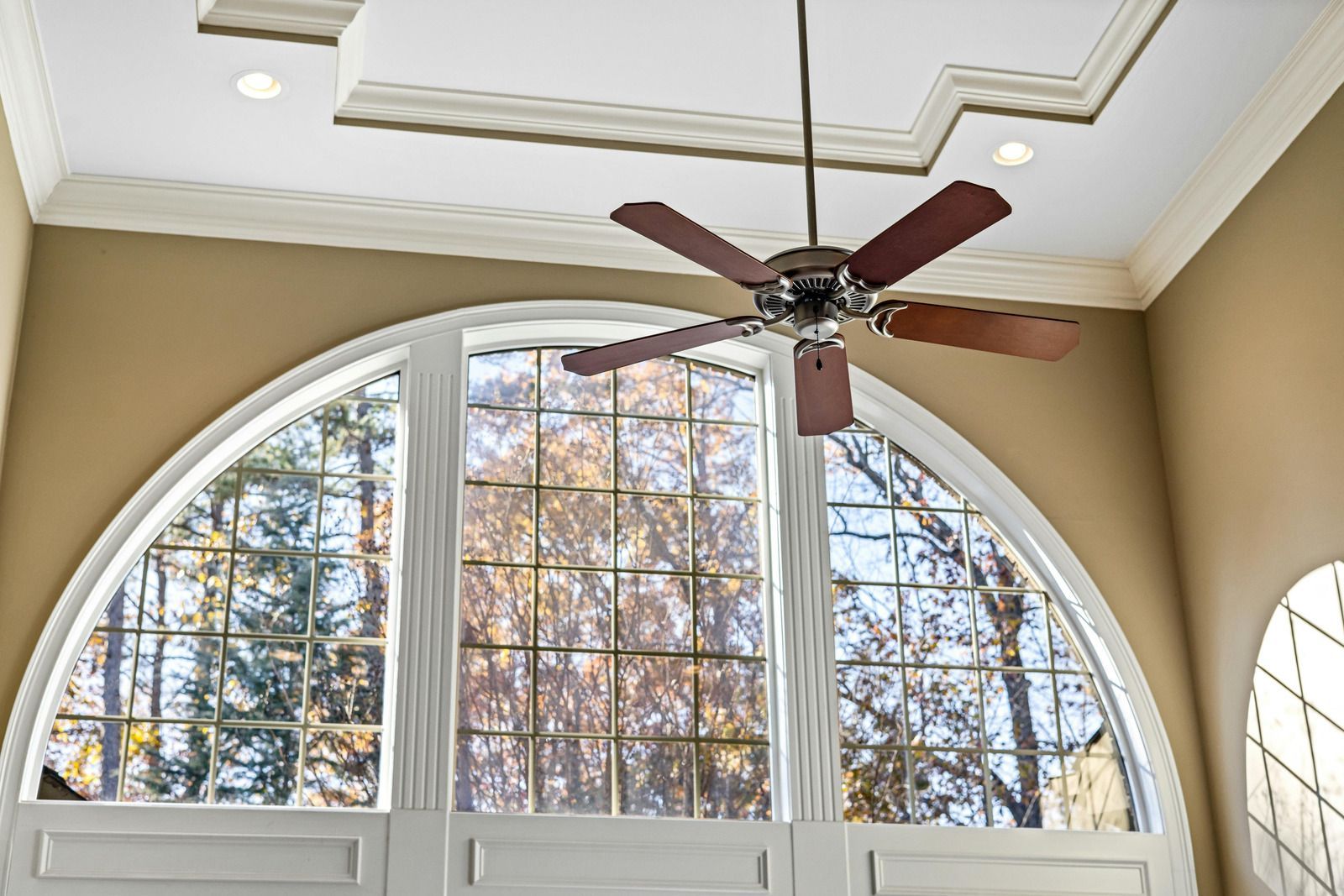 Large arched window with grids, tan walls, ceiling fan, and fall trees outside.