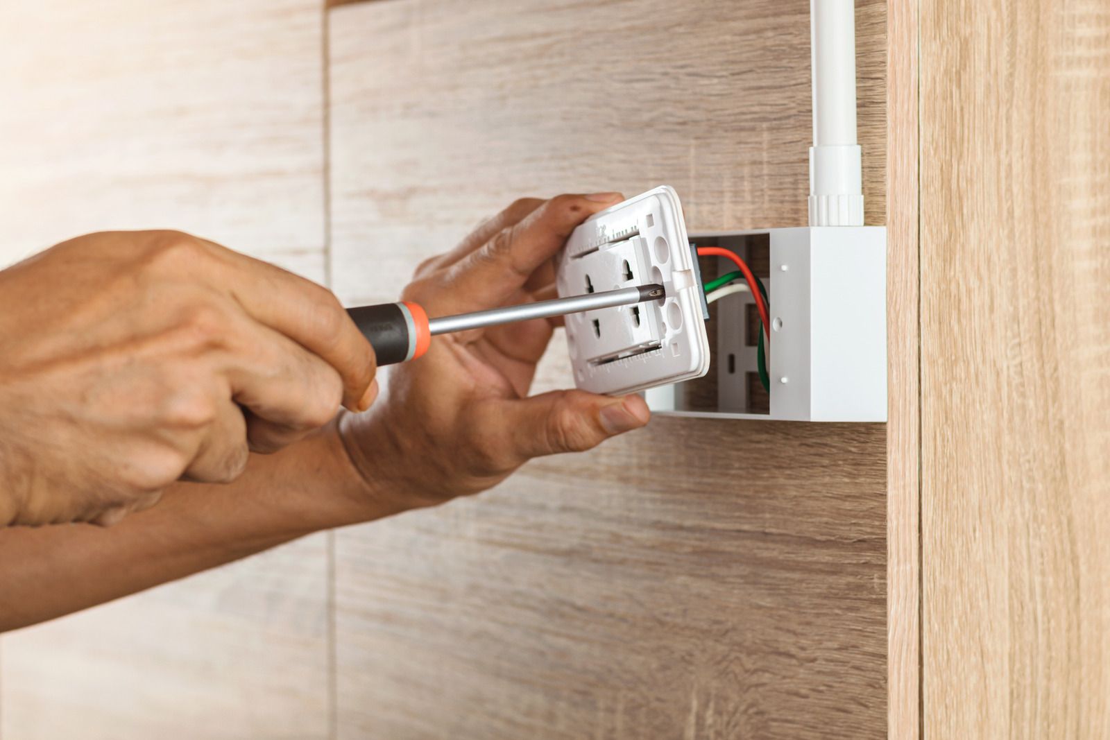 Person installing a white electrical outlet on a wooden wall using a screwdriver.