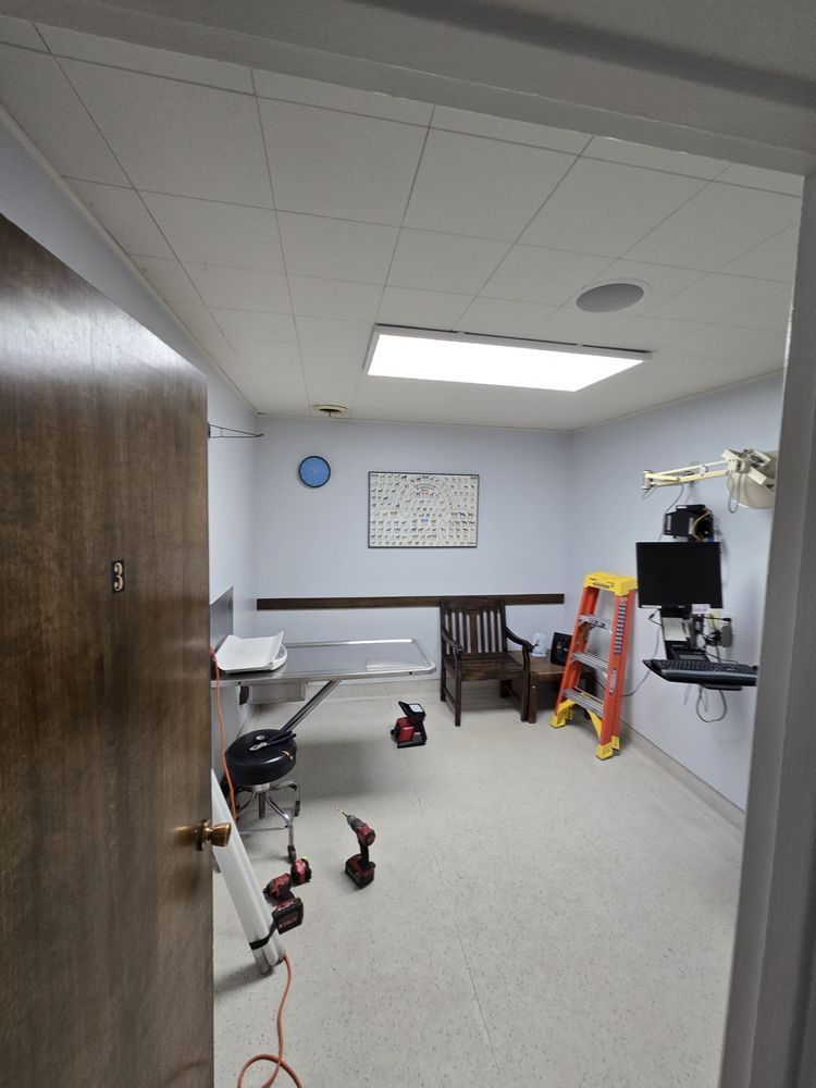 Room with exam table, chair, and equipment. Light blue walls, white ceiling, and a doorway on the left.