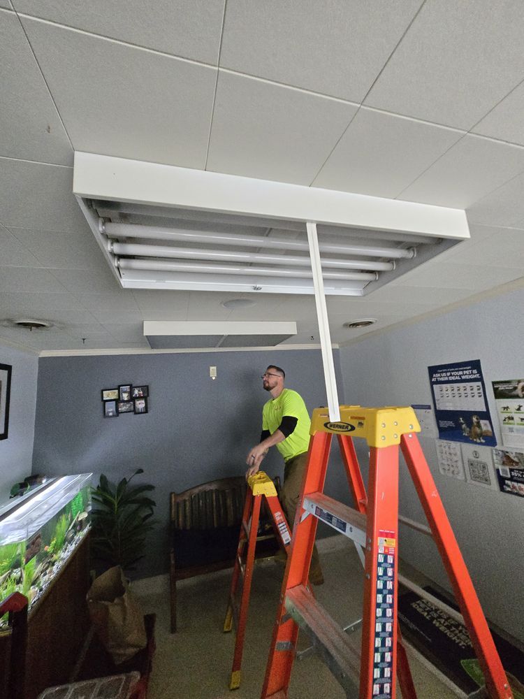 Person on a ladder repairs a fluorescent ceiling light. Interior, white ceiling, gray wall.