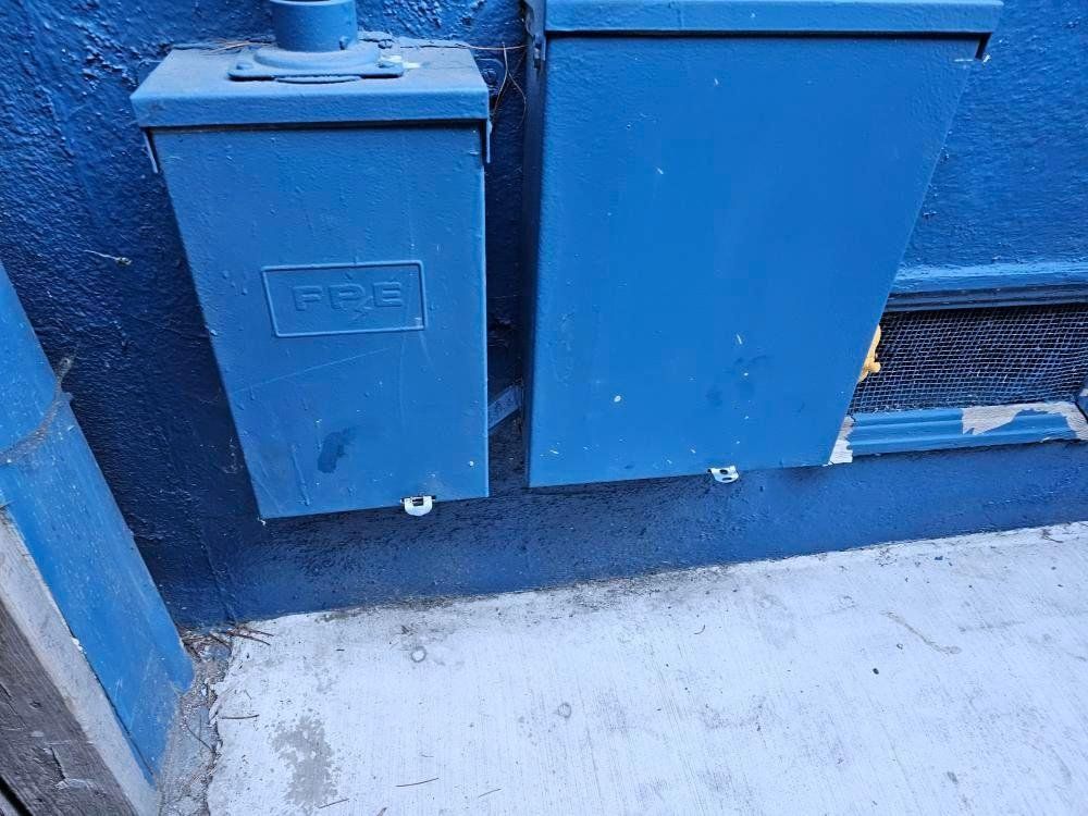 Two blue metal electrical boxes mounted on a blue wall next to a concrete surface.