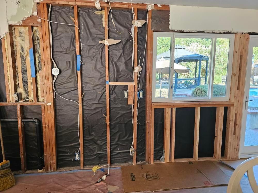 Interior wall under renovation, exposed wooden studs, wiring, window overlooking a backyard with pool.