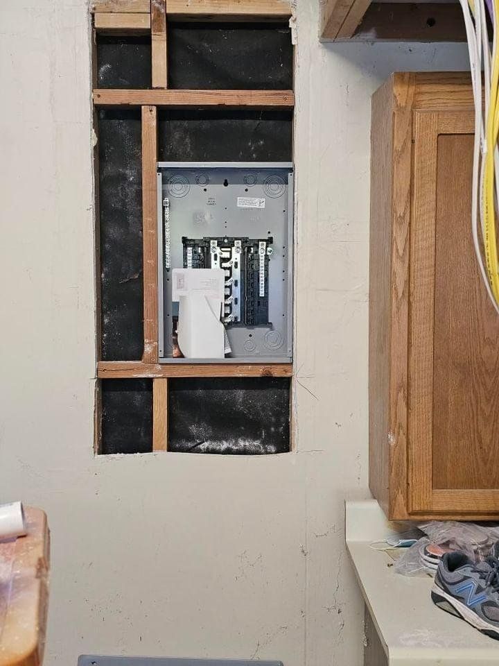 Electrical panel in a wall opening, surrounded by framing, near a kitchen cabinet.