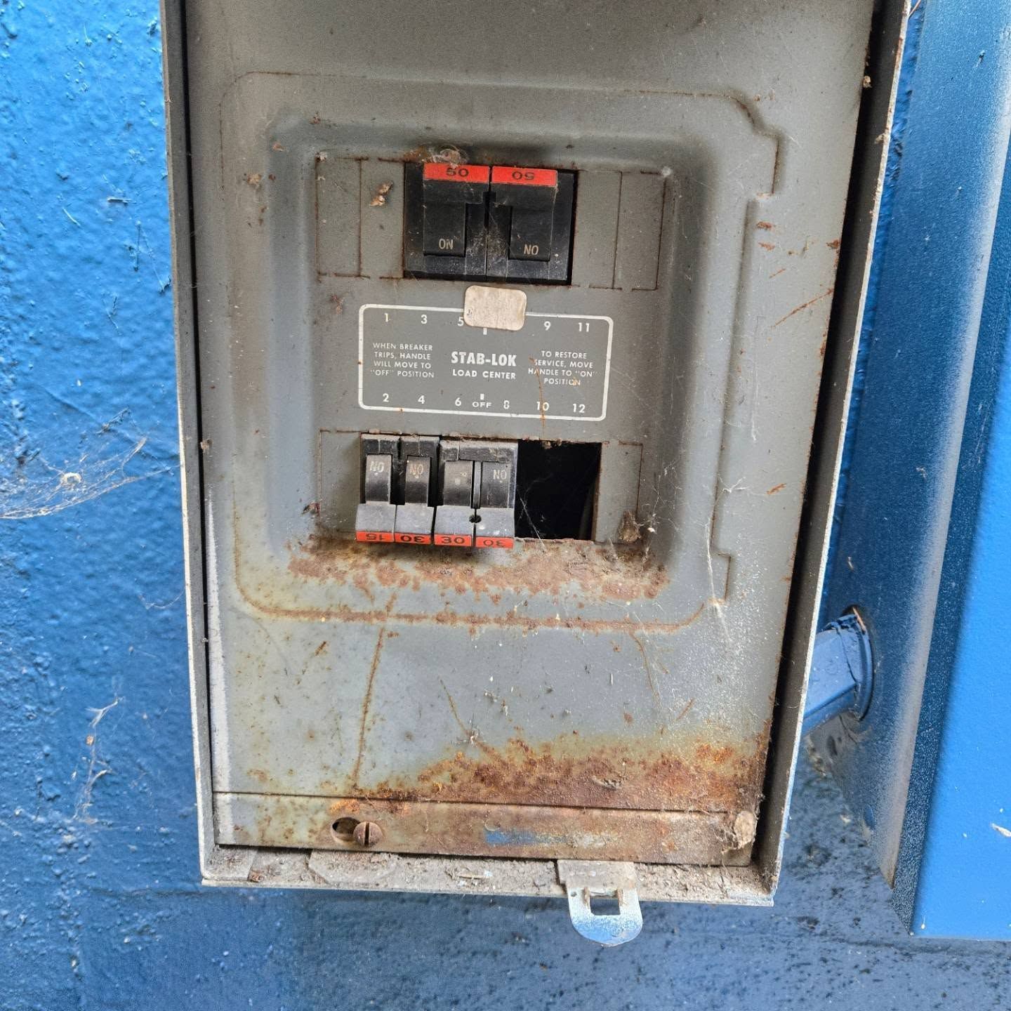 Rusty electrical breaker box mounted on a blue wall. Two breakers are on. One slot is empty.