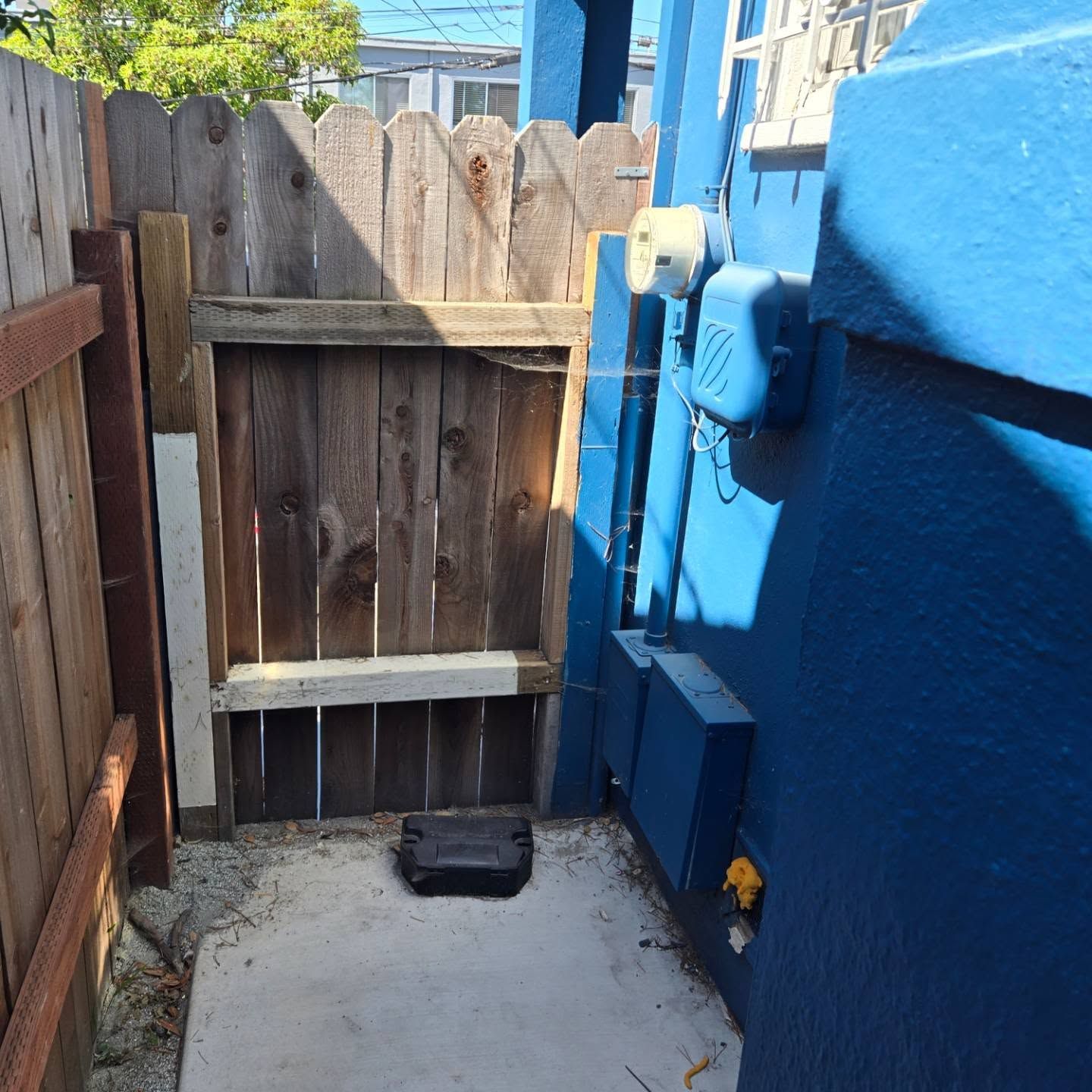 A wooden gate in a fence next to a blue building with electrical boxes and a cable.