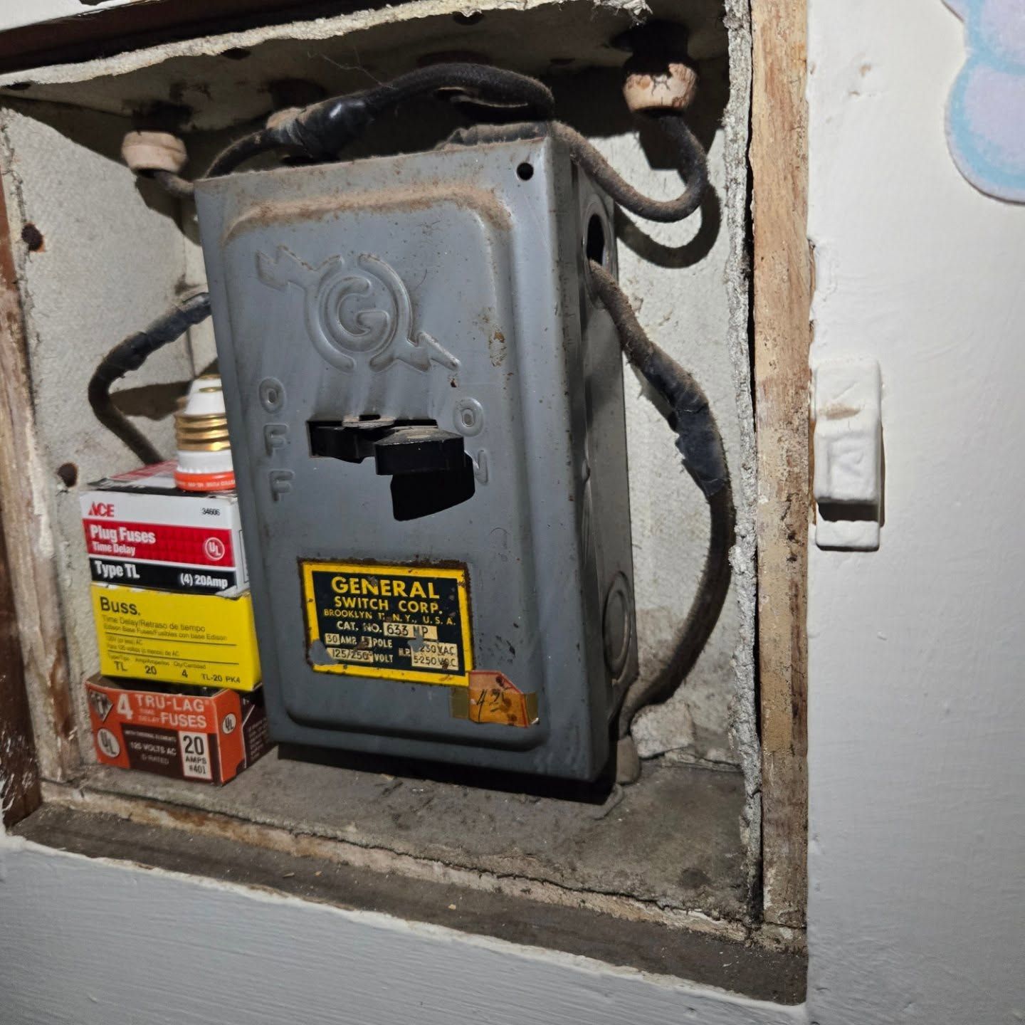 Gray electrical box with wires, in a wall, containing fuse boxes. A light switch is beside it.
