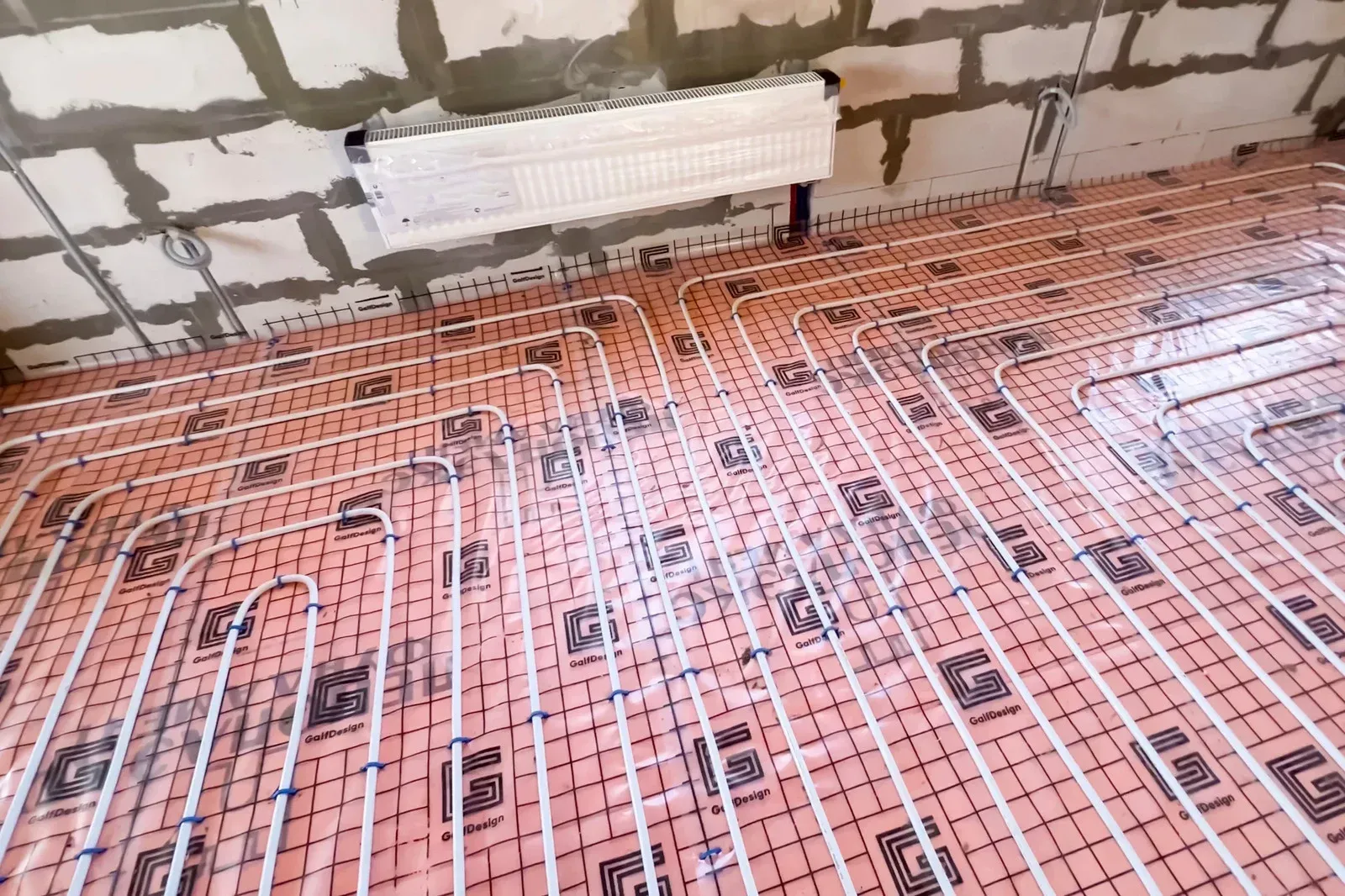 Floor heating system installation in progress. Pink insulation with white tubing, near a brick wall and a radiator.