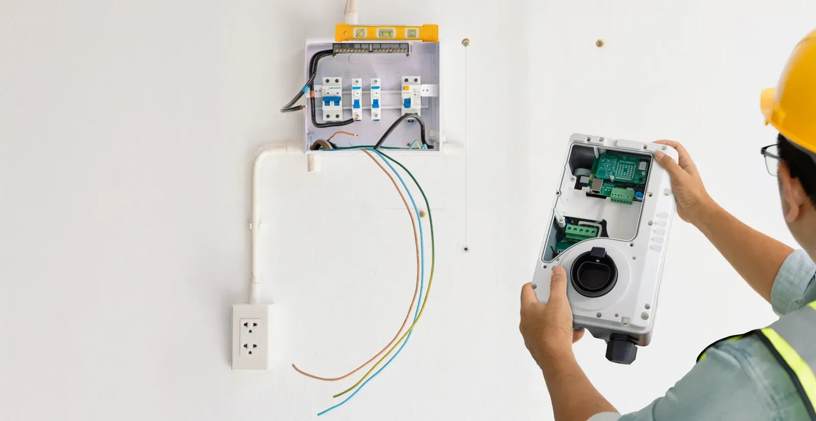 Electrician in yellow hard hat working on wiring near a breaker box and electrical outlet.
