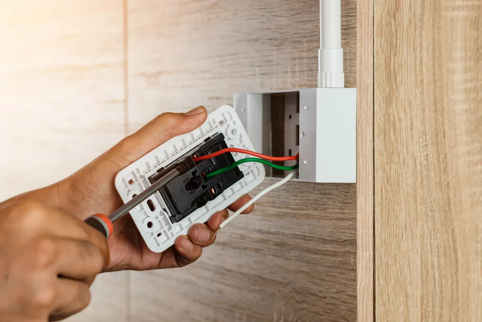 Hands fixing a white electrical outlet with a screwdriver; wires visible, wood panel background.