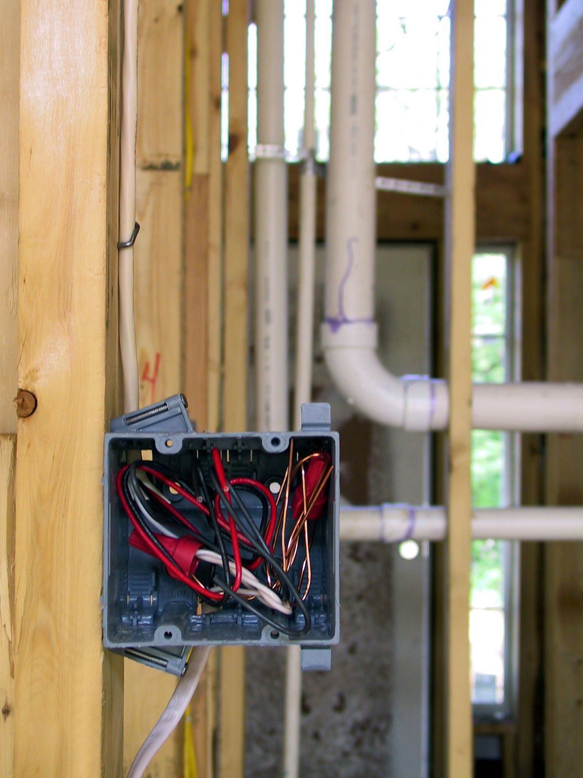 Electrical box with exposed wires attached to wooden studs, with plumbing pipes visible in the background.
