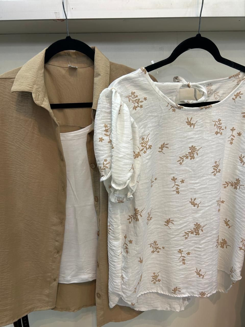 Camisa abotonada de color canela y top floral blanco colgados en perchas negras contra una pared blanca.