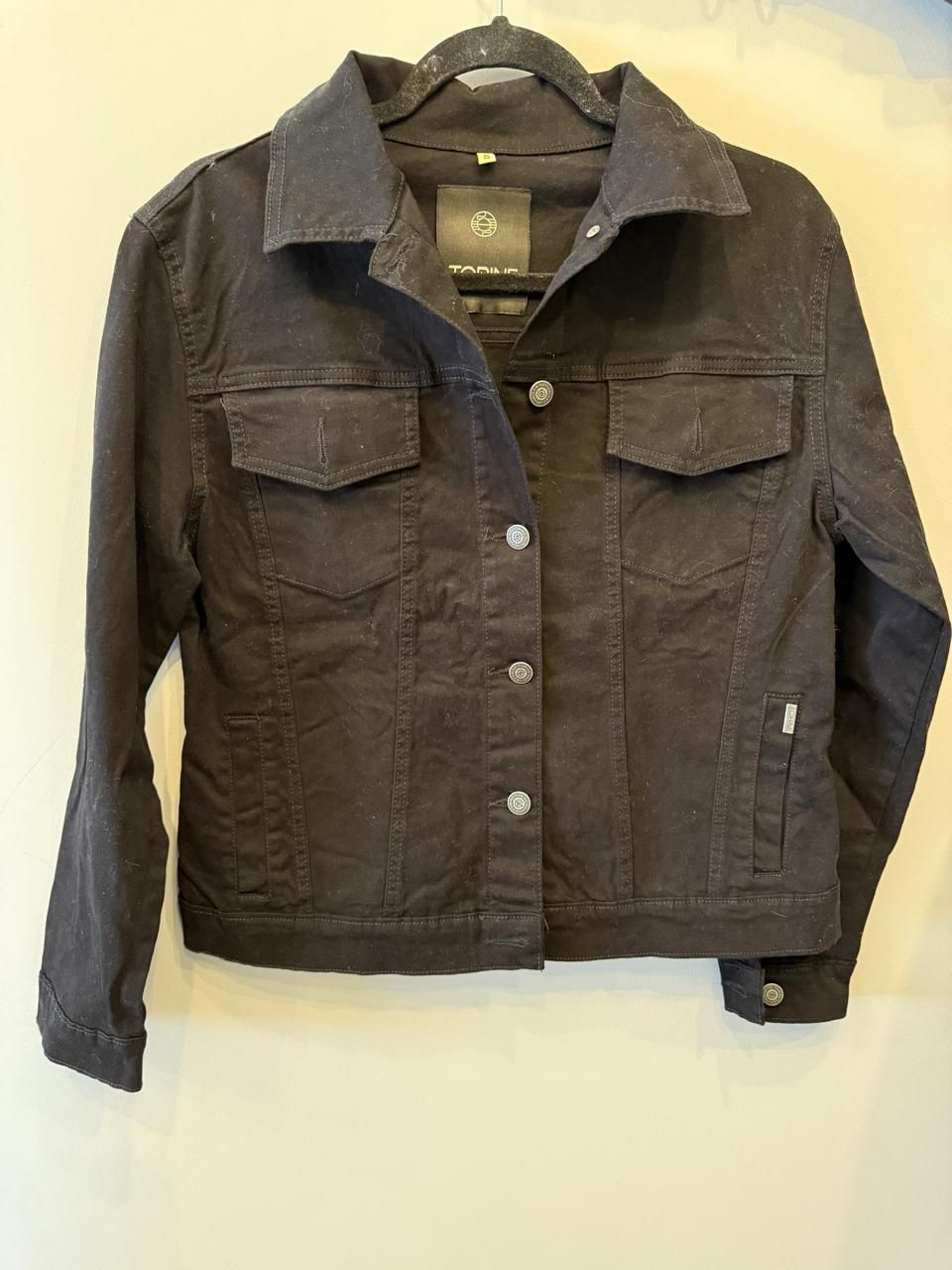 Chaqueta vaquera azul claro colgada en una percha negra, con una etiqueta adjunta.