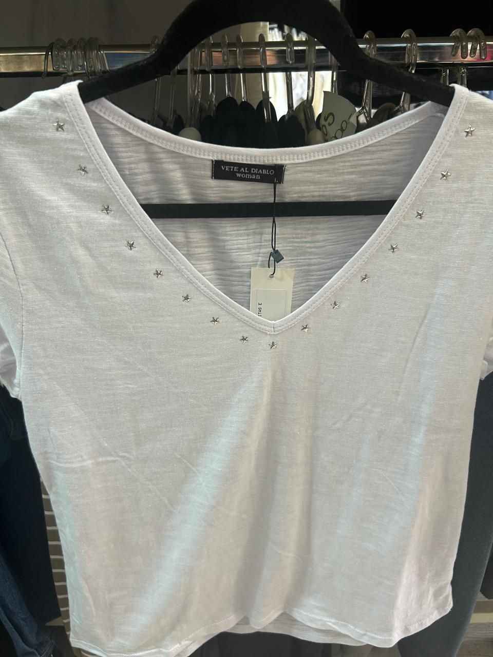 Camiseta blanca con cuello en V y pequeños tachones alrededor del escote, colgada en una percha negra.