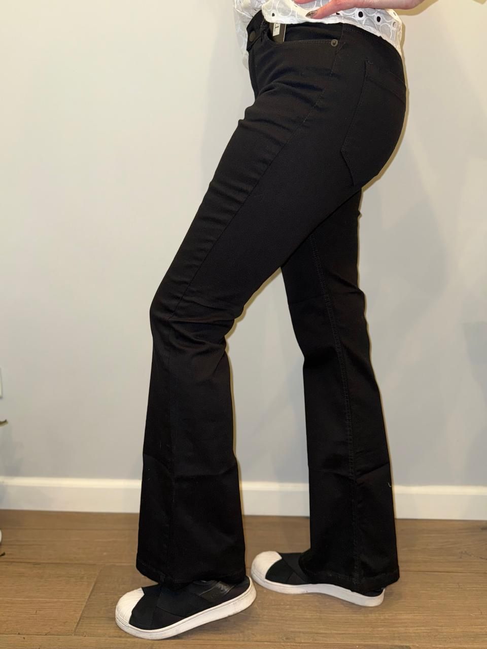 Mujer con pantalones acampanados negros y zapatillas blancas, posando en el interior.