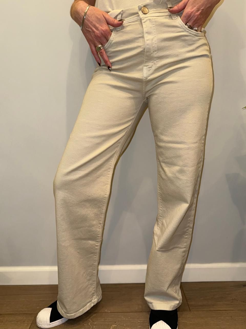 Persona con vaqueros color canela claro y zapatillas negras. De pie junto a una pared gris claro.