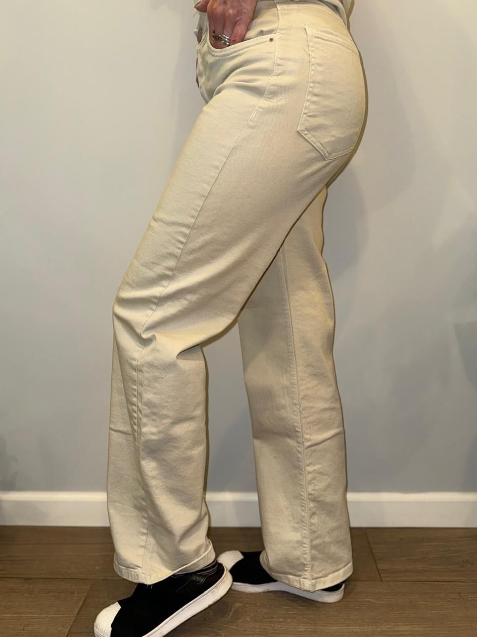 Persona con vaqueros color canela claro y zapatillas negras. De pie junto a una pared gris claro.