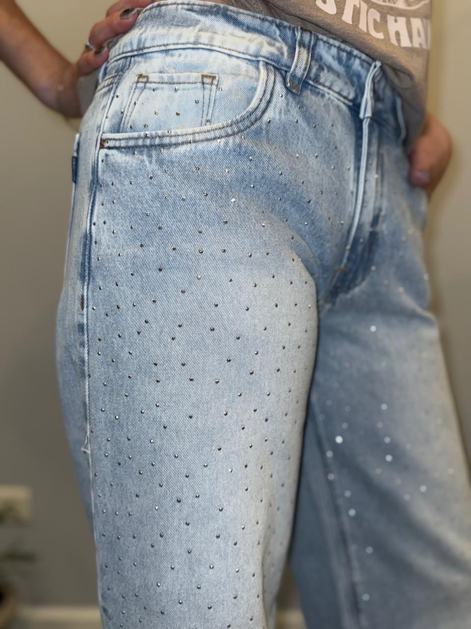 Vaqueros de lavado claro con pedrería dispersa. Persona con vaqueros, manos en los bolsillos y zapatos blancos y negros.