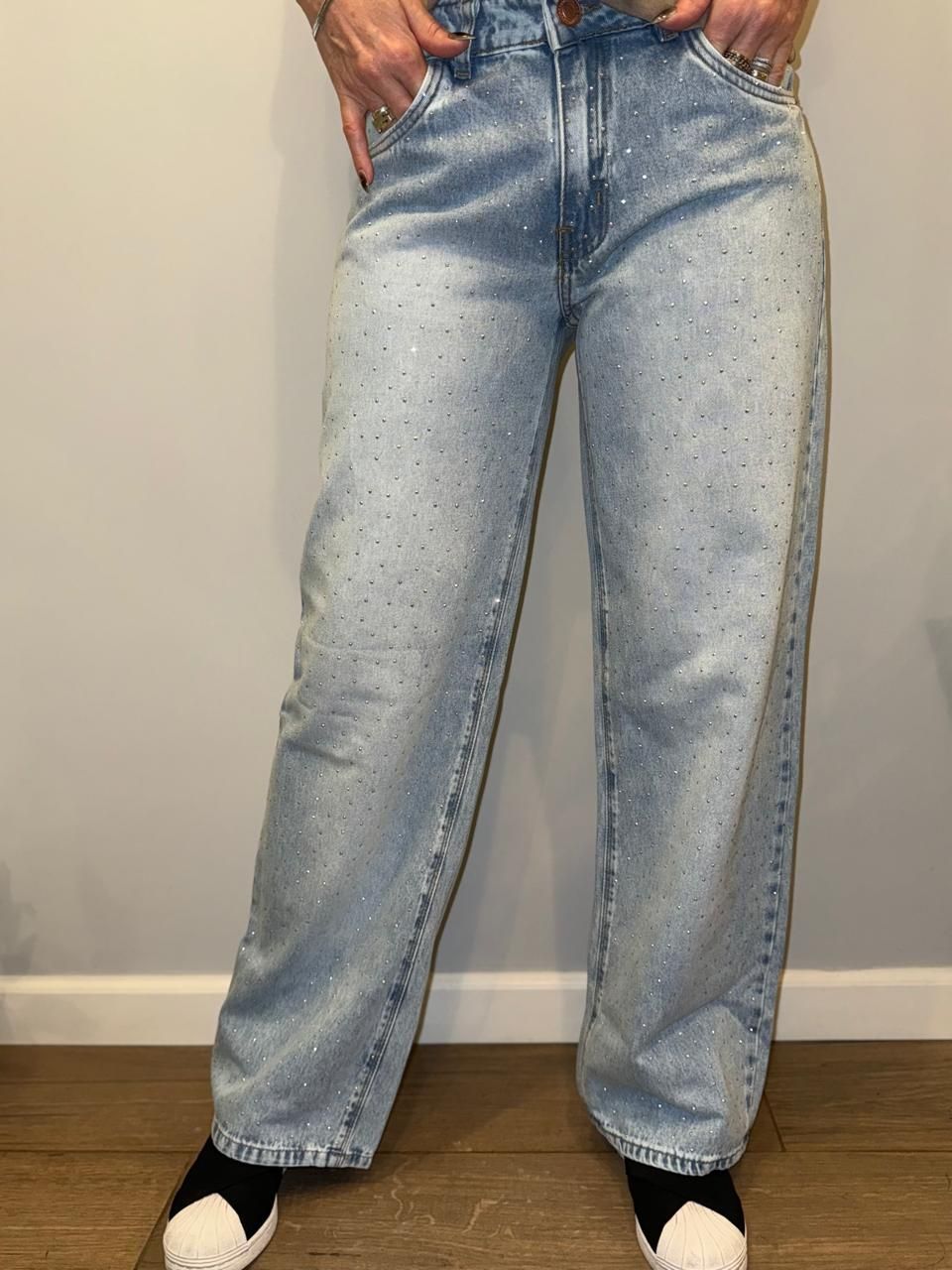 Vaqueros de lavado claro con pedrería dispersa. Persona con vaqueros, manos en los bolsillos y zapatos blancos y negros.
