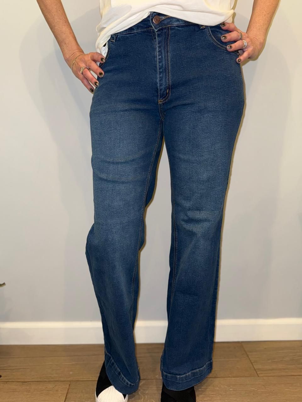 Mujer que viste pantalones vaqueros acampanados de color azul y zapatillas deportivas frente a una pared gris.