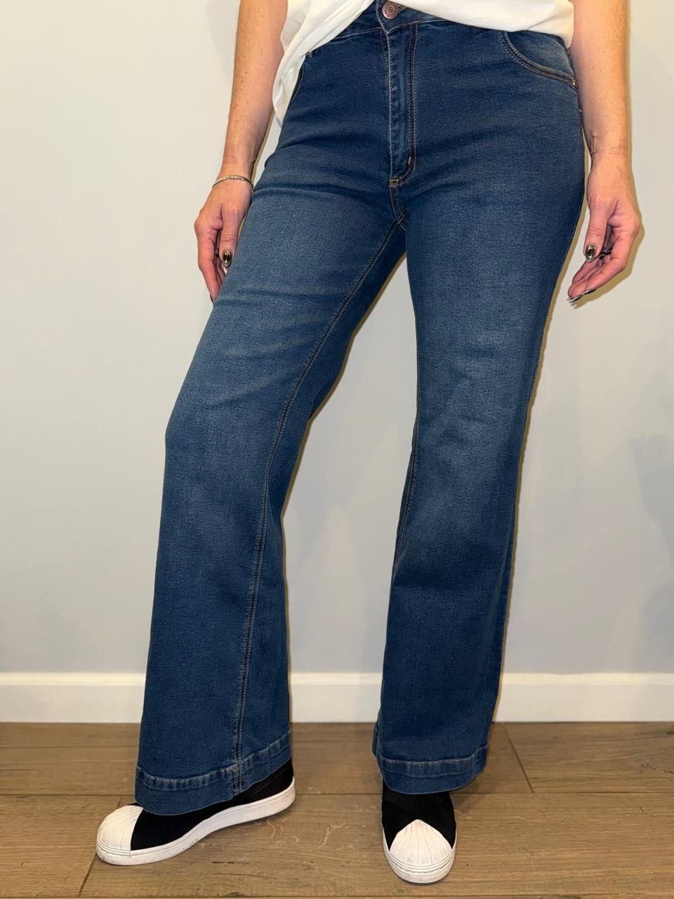 Mujer que viste pantalones vaqueros acampanados de color azul y zapatillas deportivas frente a una pared gris.