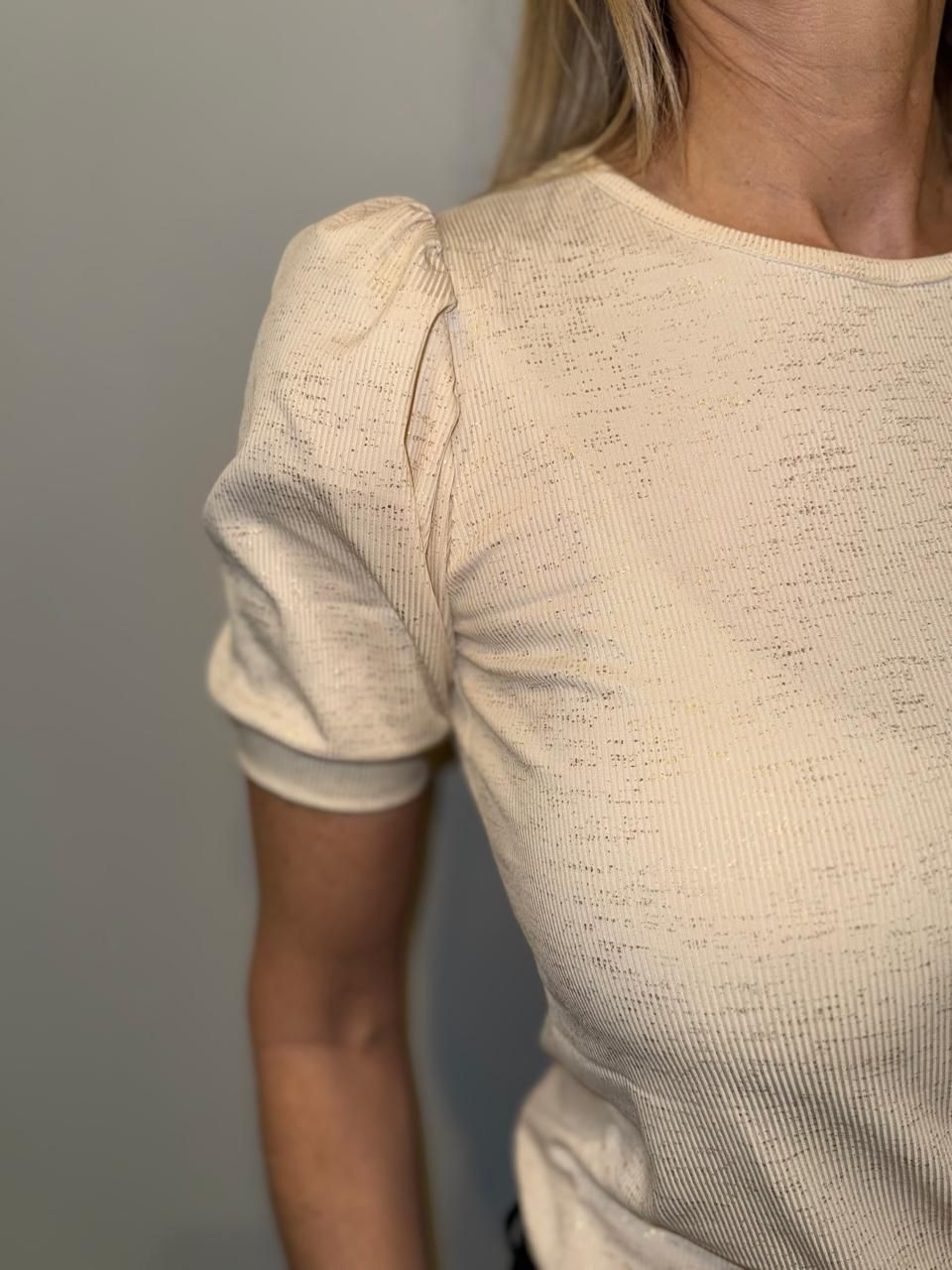 Mujer con una blusa texturizada de color crema y mangas abullonadas, de pie cerca de una pared gris.