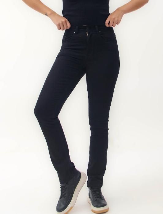 Mujer que viste jeans negros y zapatillas de deporte, de pie sobre un fondo blanco, subiendo la cintura de los jeans.