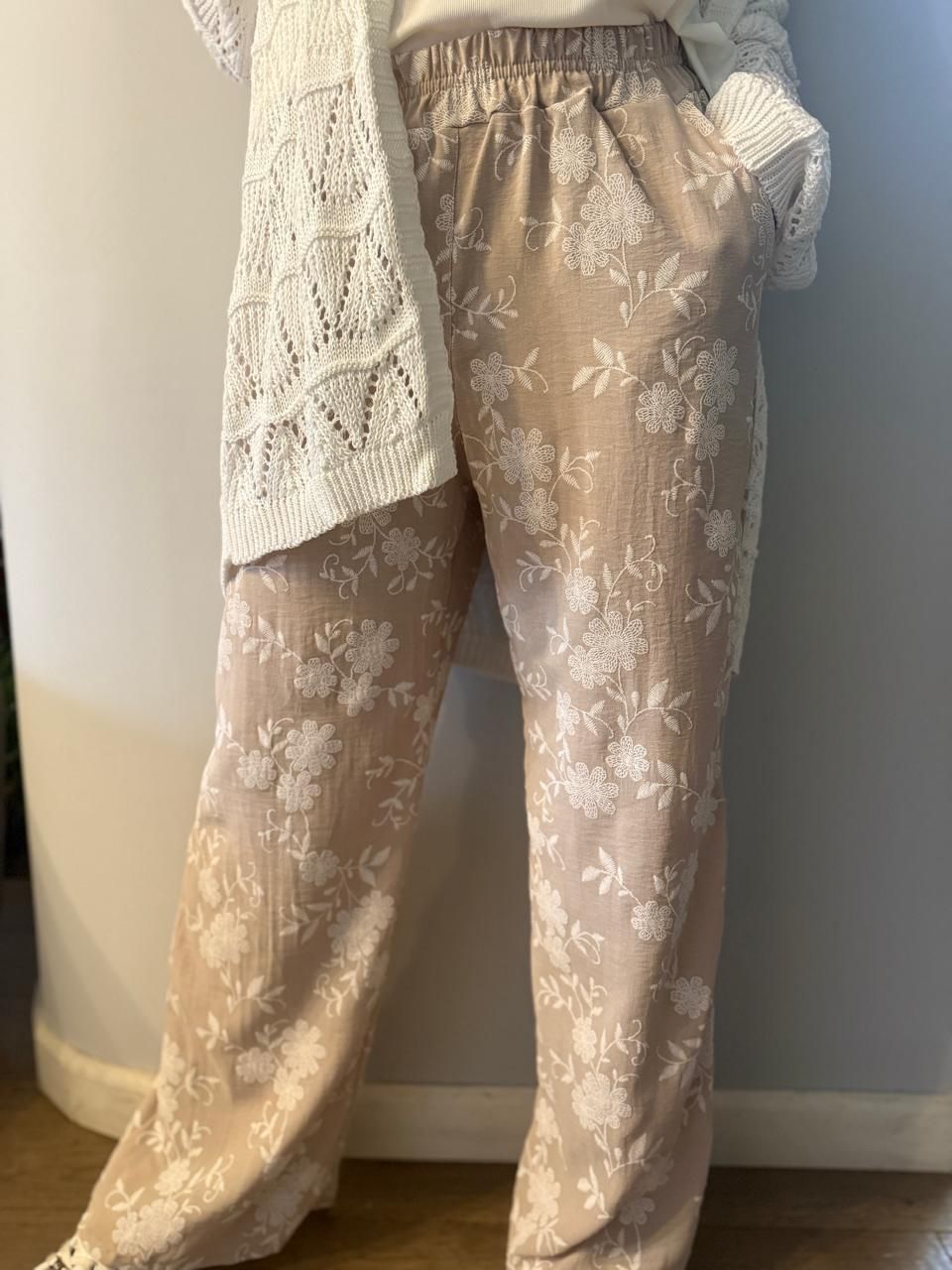 Persona que viste pantalones beige con estampado floral y suéter blanco, de pie en el interior.