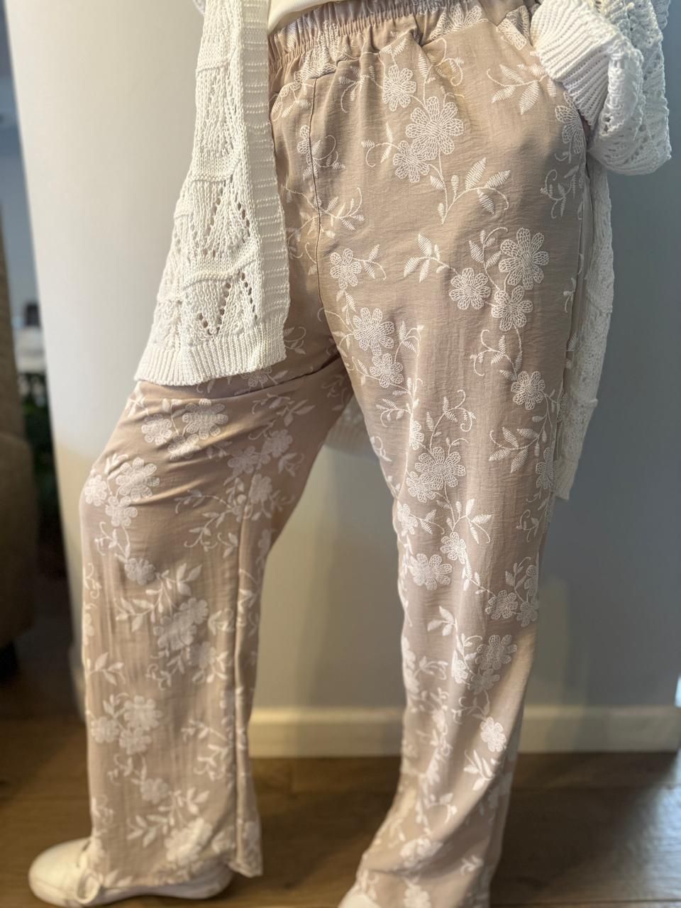 Persona que viste pantalones beige con estampado floral y suéter blanco, de pie en el interior.
