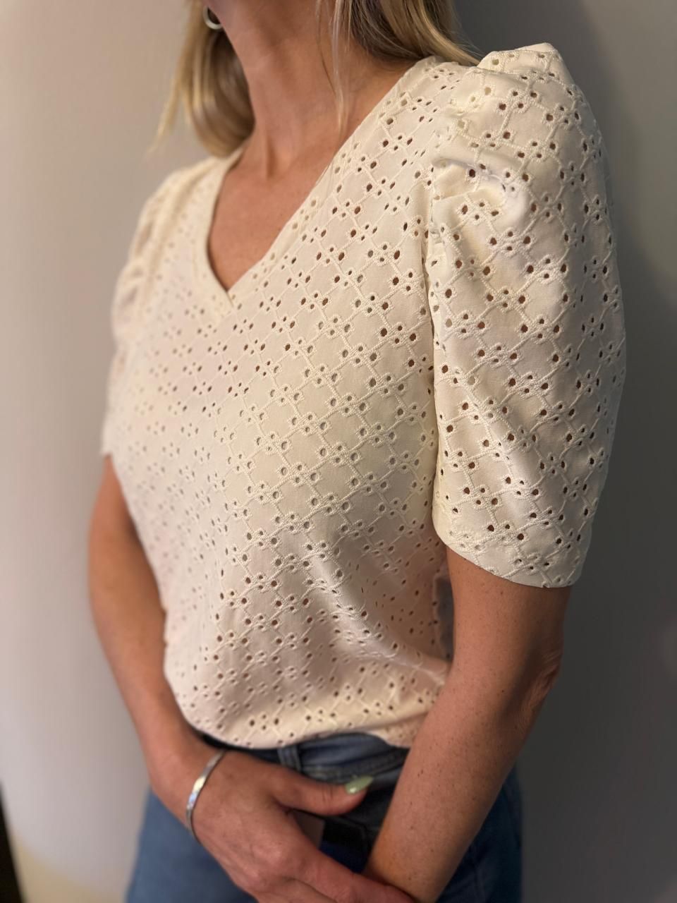 Mujer con un top de escote en V con ojales de color blanco roto y mangas abullonadas, vestida con jeans azules.