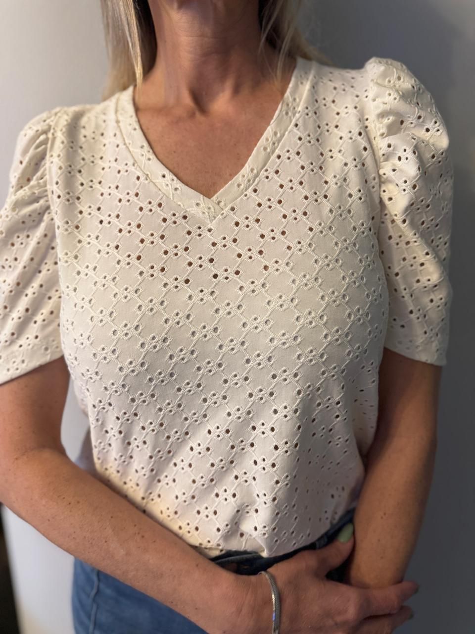 Mujer con un top de escote en V con ojales de color blanco roto y mangas abullonadas, vestida con jeans azules.