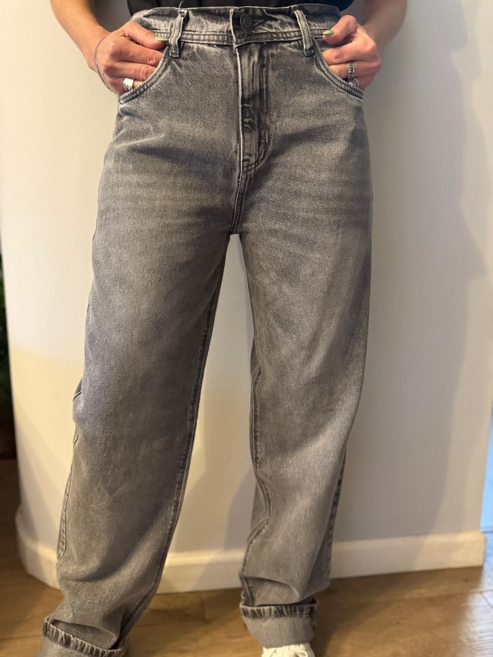 Persona con vaqueros grises deslavados, con las manos en los bolsillos, apoyada contra una pared blanca.