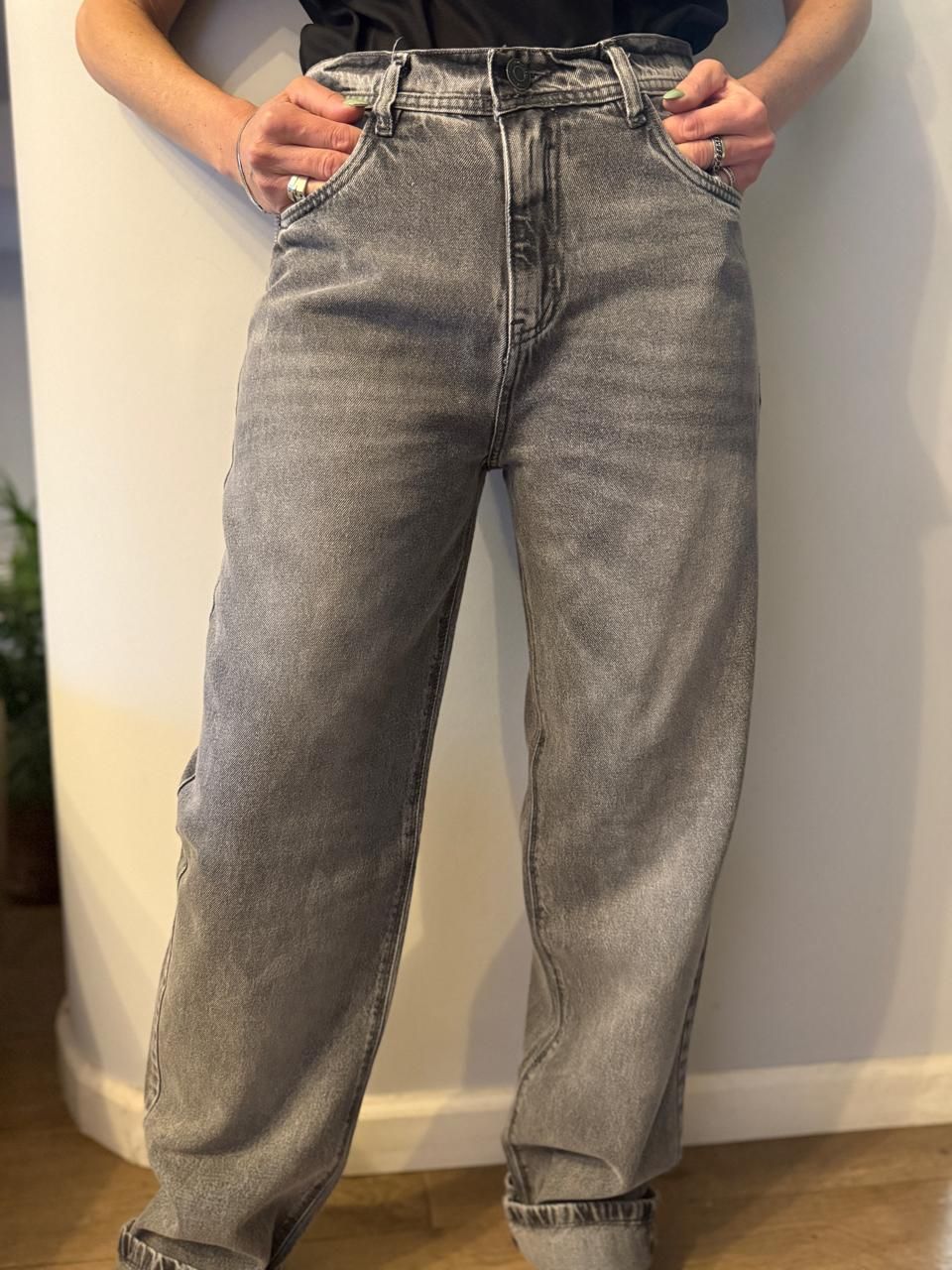 Persona con vaqueros grises deslavados, con las manos en los bolsillos, apoyada contra una pared blanca.