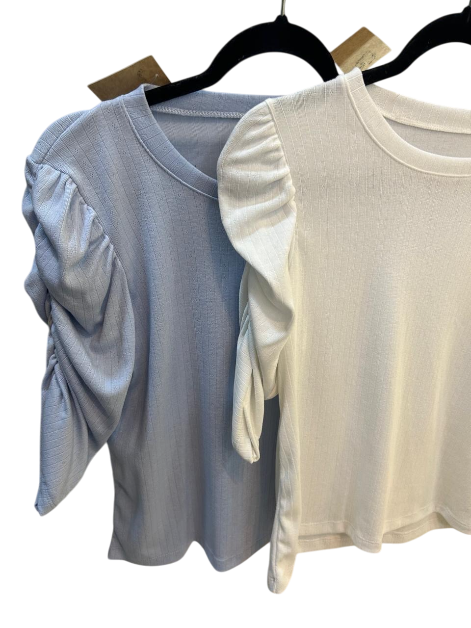 Dos blusas de manga corta con mangas abullonadas, una azul claro y otra blanca, colgadas en perchas negras.