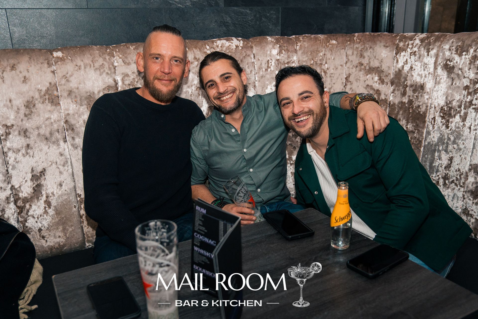 Three men are posing for a picture while sitting at a table in a bar.
