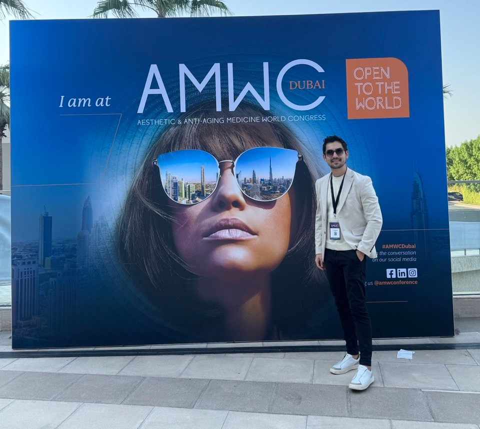 Homem de blazer e óculos escuros em pé ao lado de um painel de fundo do evento AMWC Dubai.