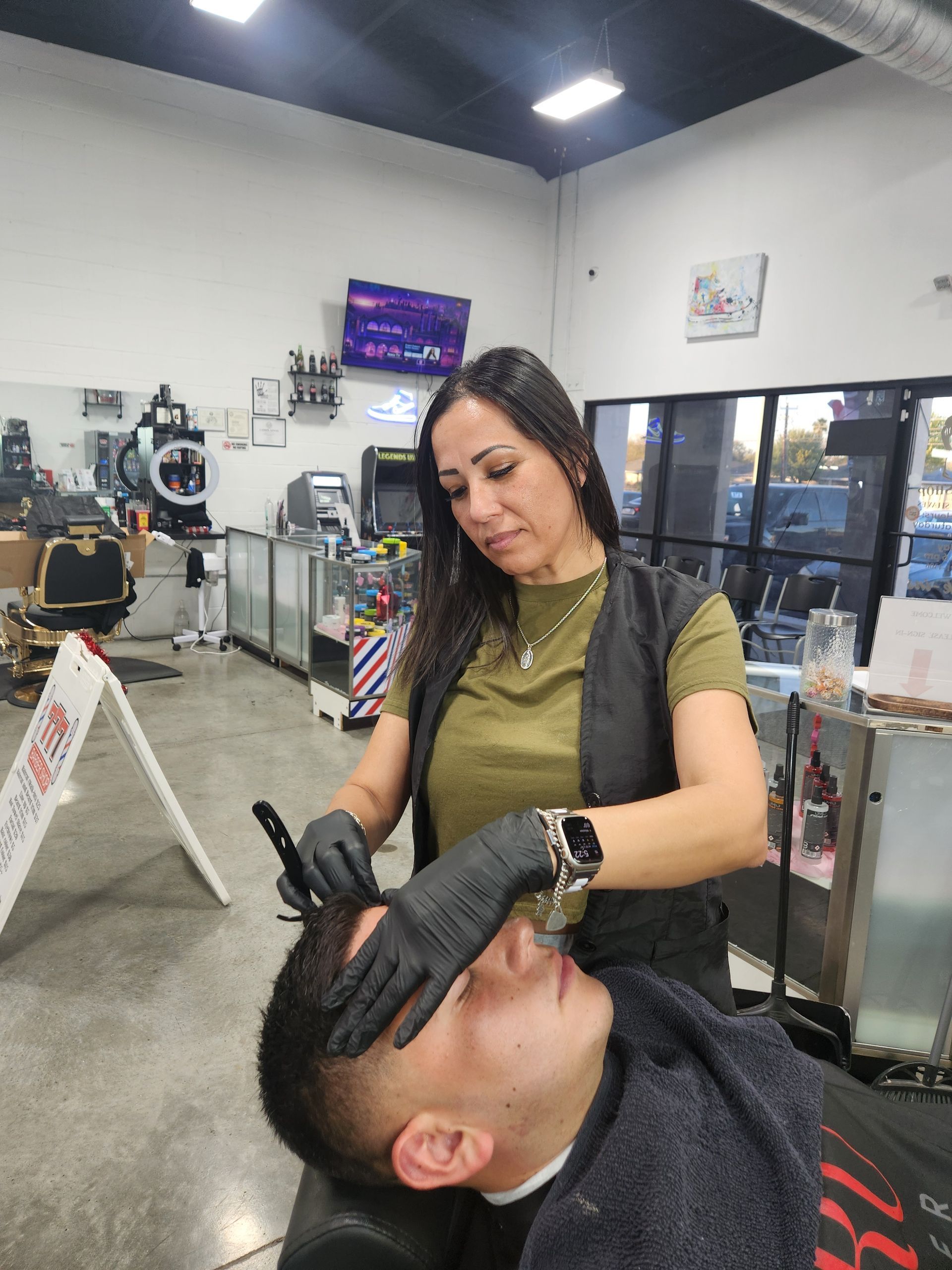 Barber giving a customer a haircut in a shop. Woman with black gloves, olive shirt. Man in chair.
