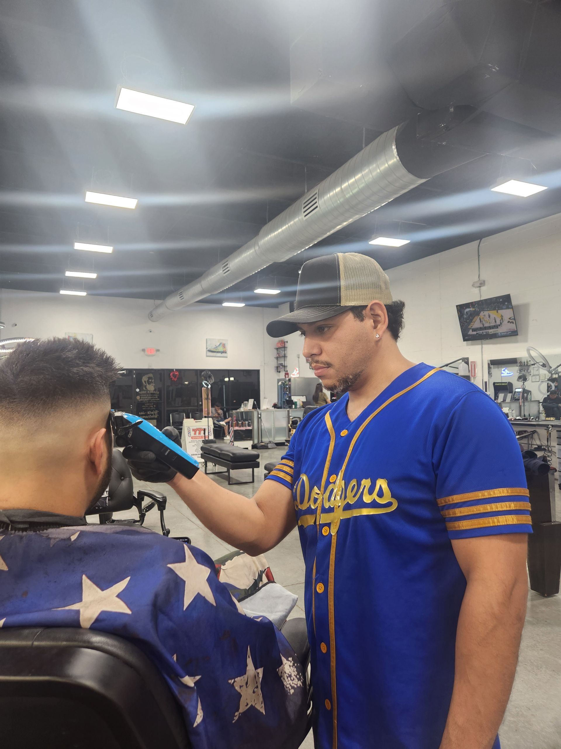 A barber in a Dodgers jersey cutting a client's hair in a shop. The client is seated and draped.