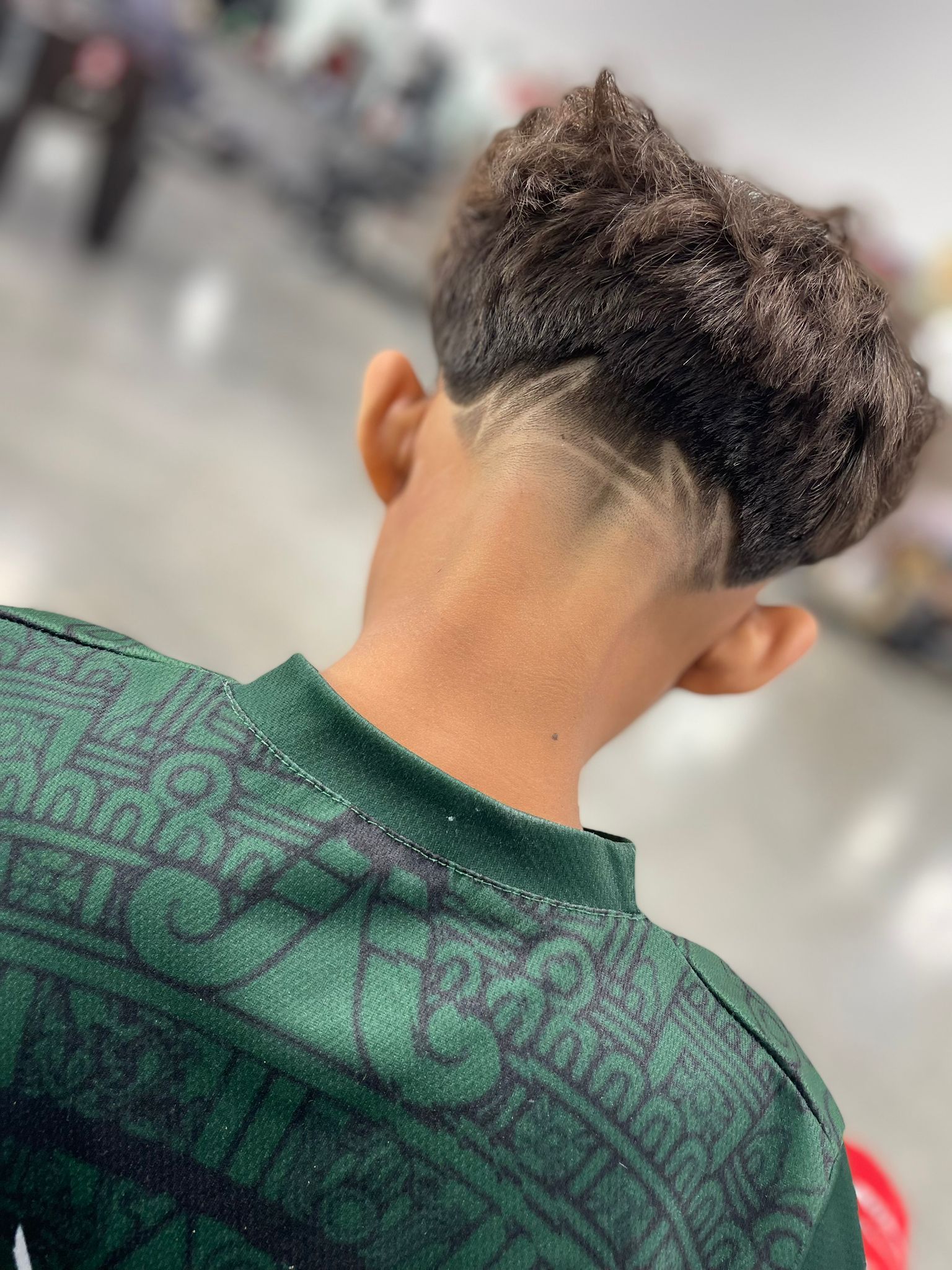 Person's back, wearing a green sweater, with a high top haircut and shaved design. Indoors.