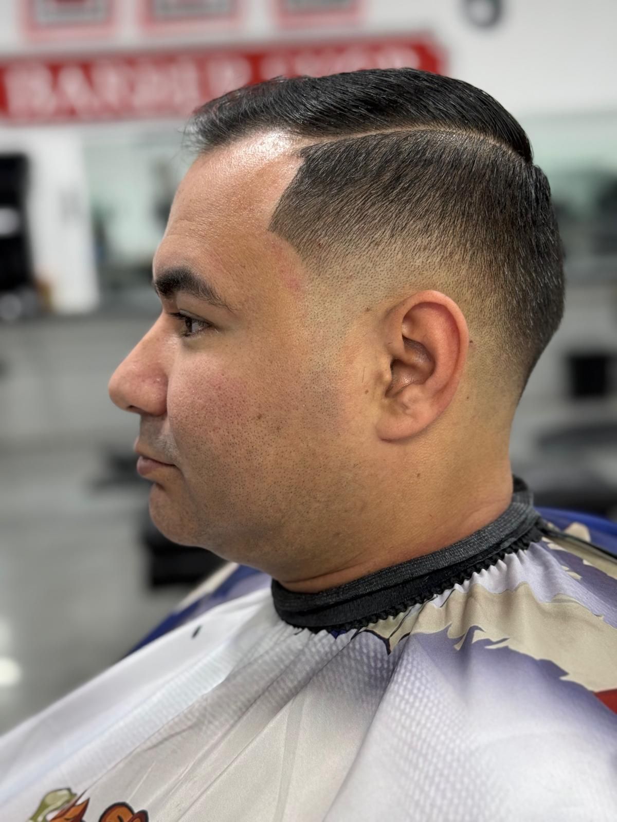 Man with a fade haircut and side part, wearing a barber cape, in a barber shop.