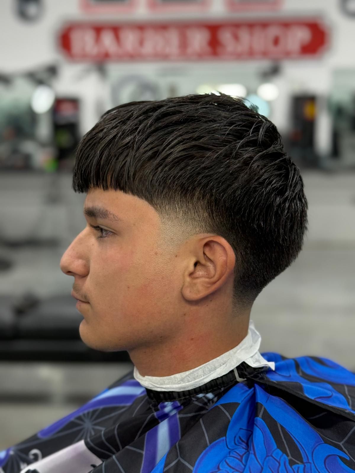 Man with a high fade haircut in a barber shop, wearing a blue cape.