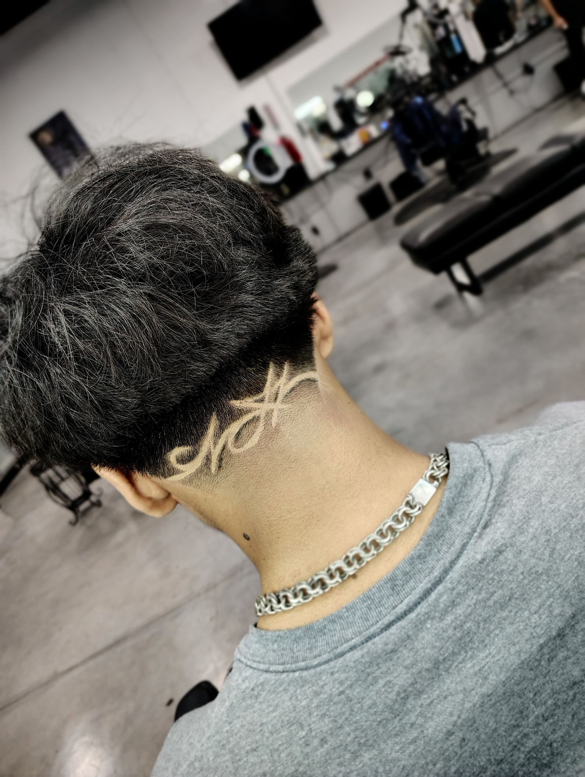 Person's nape with textured dark hair and a detailed, artistic hair design. They wear a silver chain and grey shirt.