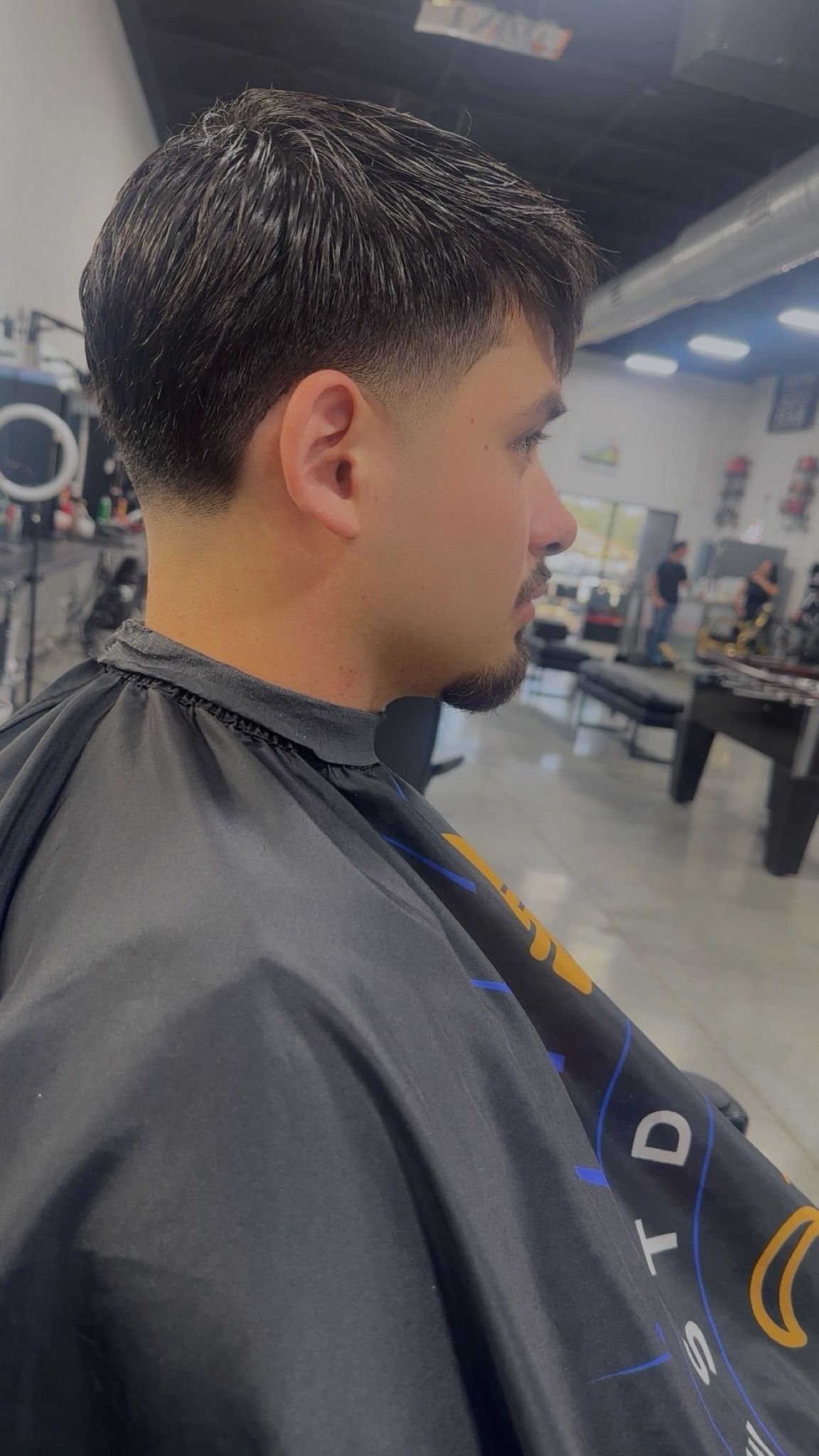 Man getting a haircut at a barbershop. He has dark hair, a low fade, and a beard. He wears a salon cape.
