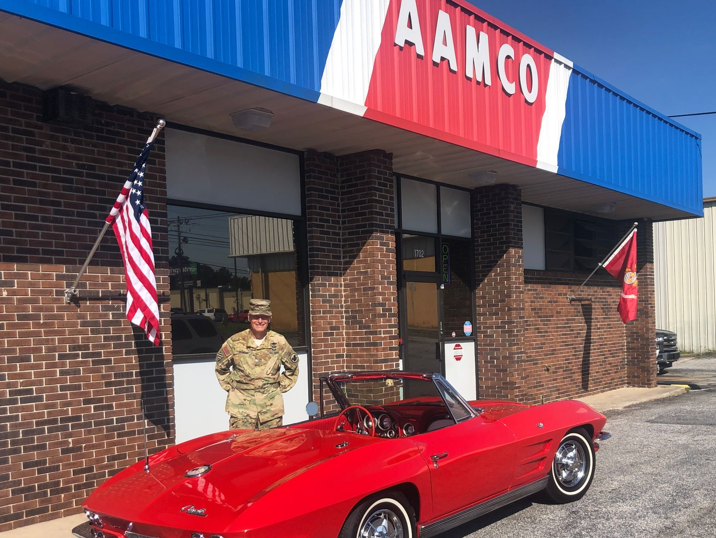 Bright red vintage convertible parked outside a brick AAMCO shop, with a person standing by the entrance.