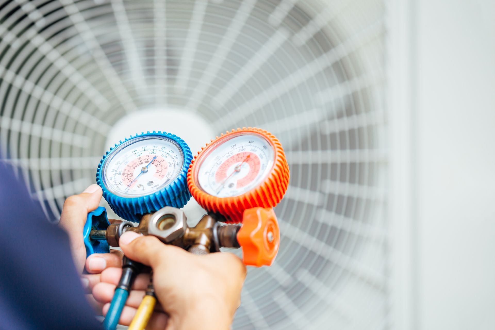 Hands holding a pressure gauge manifold with blue and red dials in front of a large fan grille