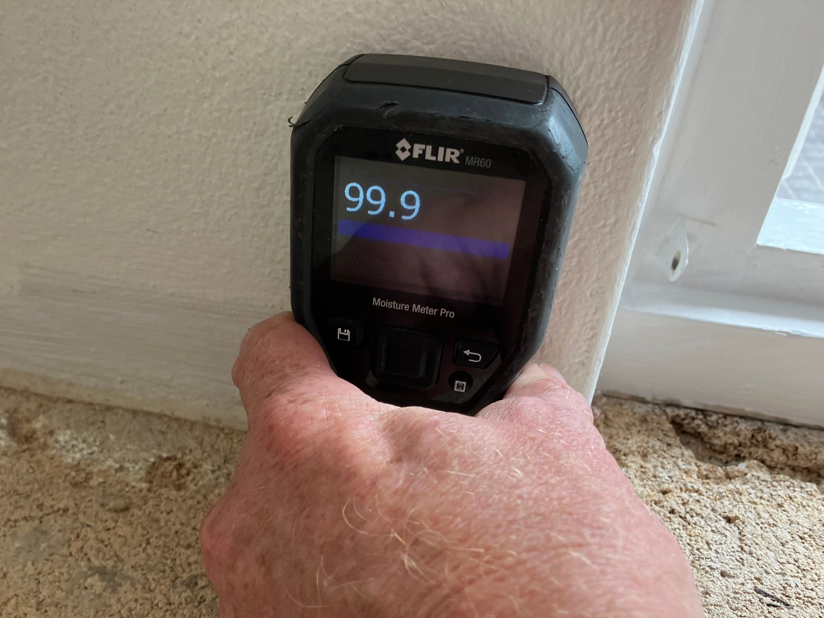 Hand holding a FLIR thermal camera, reading 99.9, on white wall near window frame.