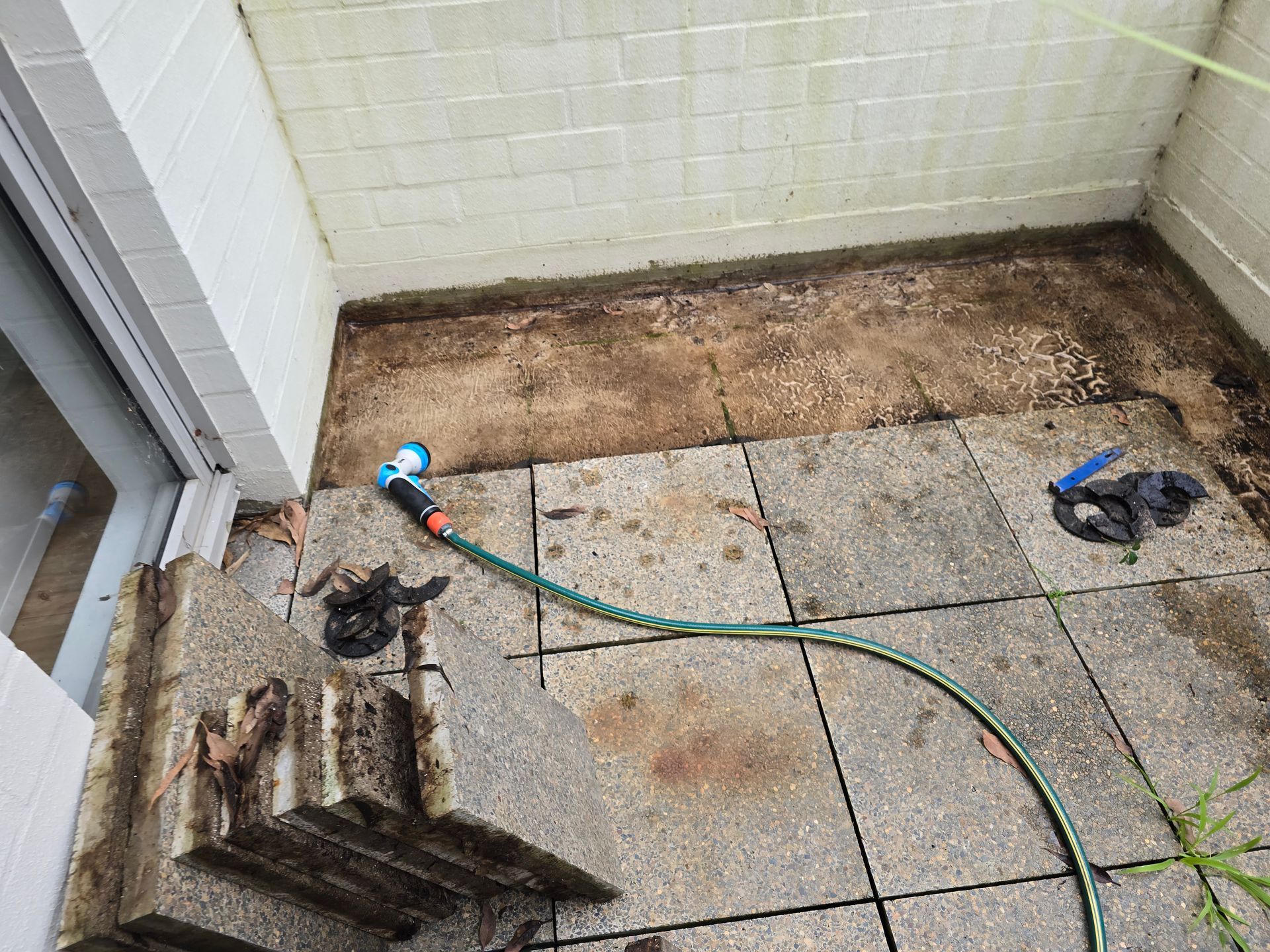 A patio with dirty stone tiles, hose, and a partially open door. Brown dirt against the walls.