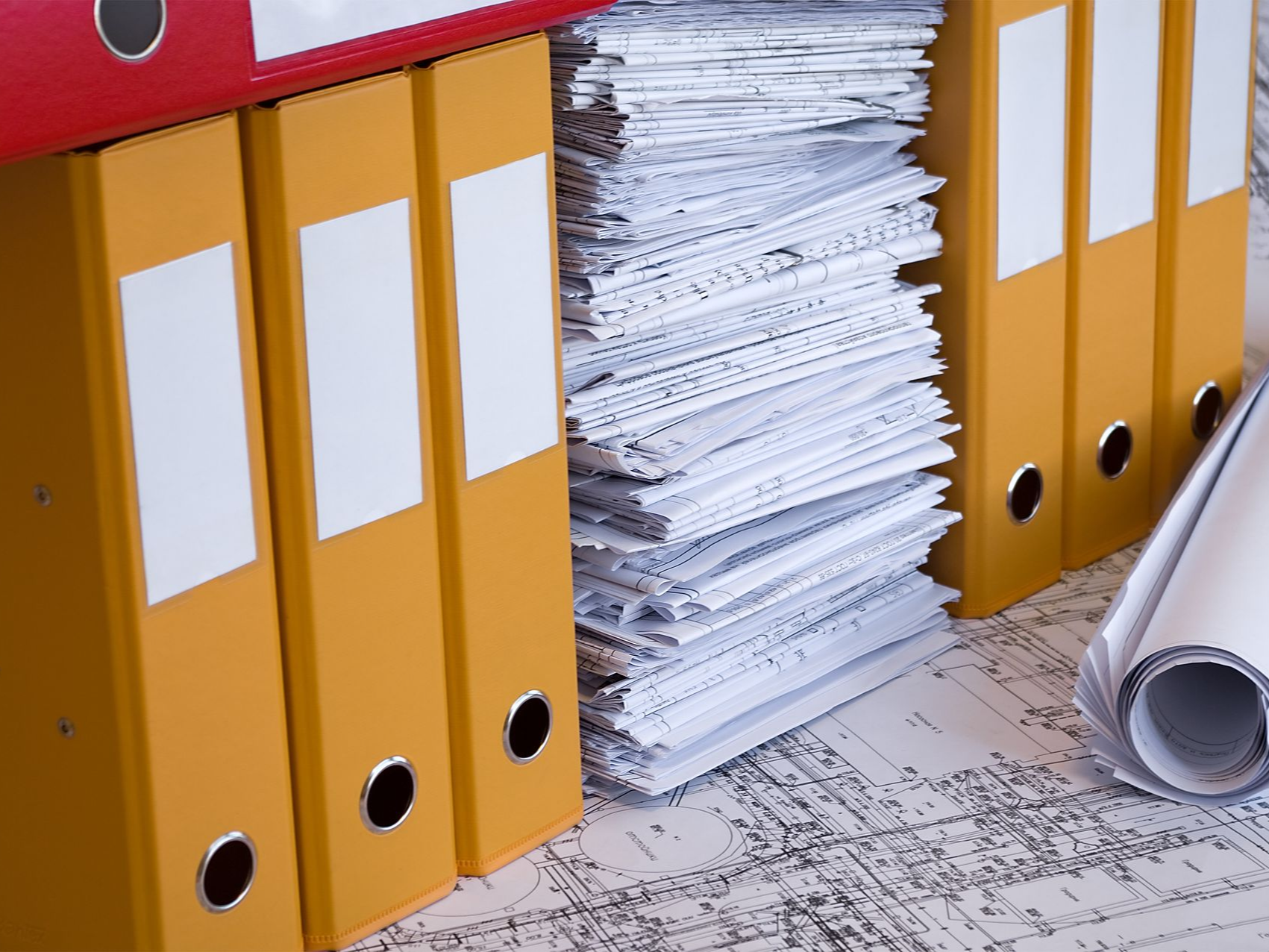 Yellow and red binders with a large stack of papers and rolled blueprints on a printed surface.