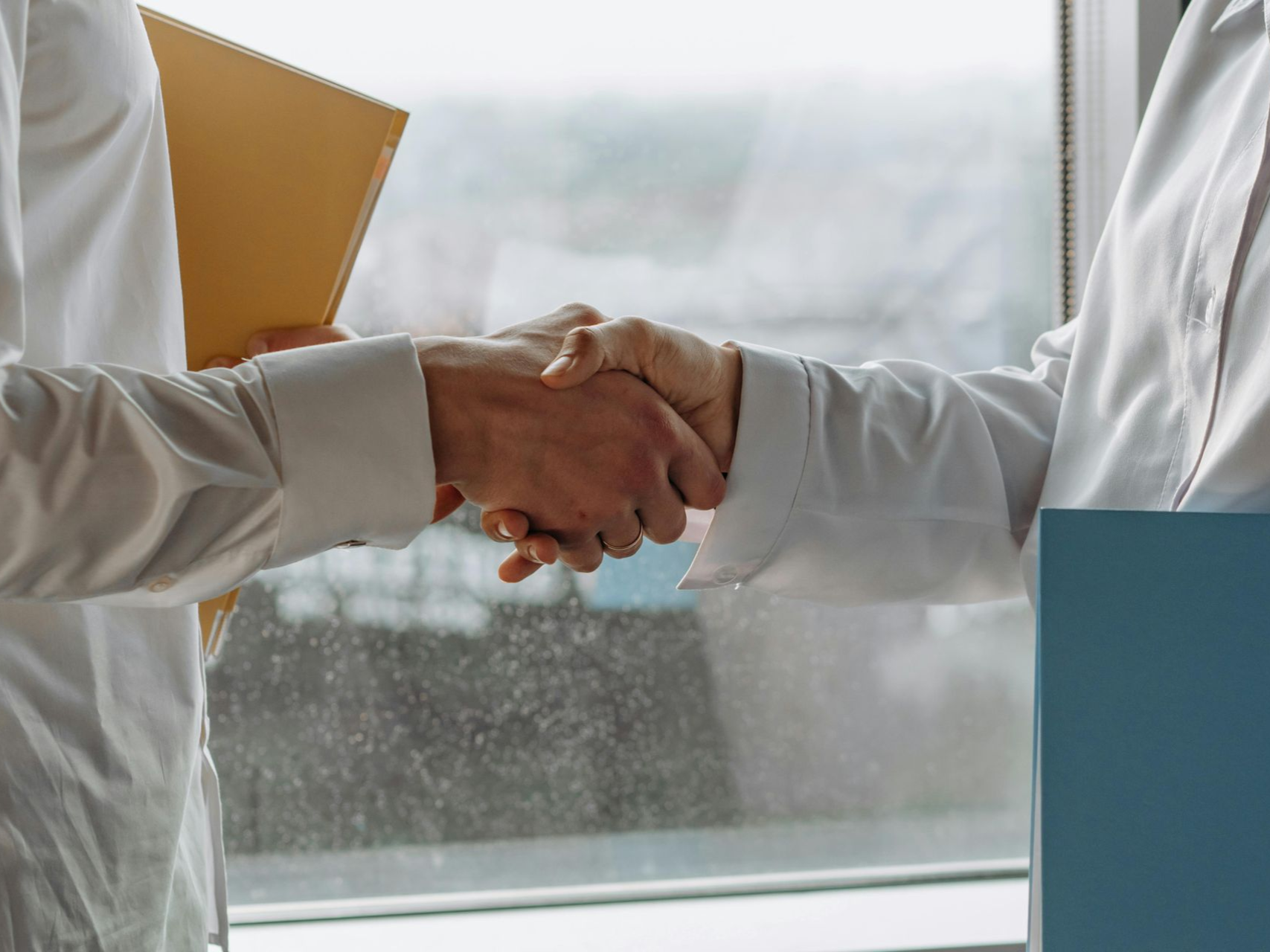 Two people shaking hands near a window, likely in an office setting. One person holds a folder.