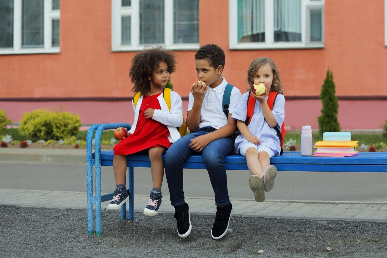 3 young children sitting outside on a bench and eating snacks