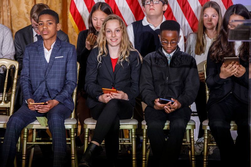 Group of people seated, some holding phones, in front of US flag.